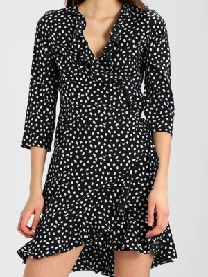 Svart omlottklänning med vita prickar - Svart kort omlottklänning med vita prickar och volangdetaljer. Klänningen har trekvartsärm och v-ringning, vilket ger en trendig och ungdomlig look. Perfekt för dig som gillar mönstrade plagg med snygga detaljer.