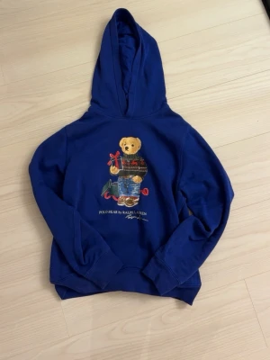 Blå hoodie Polo Ralph Lauren björn - Blå hoodie från Polo Ralph Lauren med ikoniskt björntryck på bröstet. Björnen har på sig en julig tröja och jeans, och håller ett paket. Hoodien har huva och ribbade muddar. Perfekt för dig som gillar klassisk streetstyle med en lekfull twist.