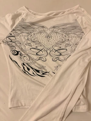 Vit långärmad topp med hjärtmönster - Vit långärmad topp med svart grafiskt tryck på ryggen som föreställer ett hjärta och vingar i en detaljerad, snirklig design. Tunn och mjuk, perfekt att styla med jeans eller kjol.
