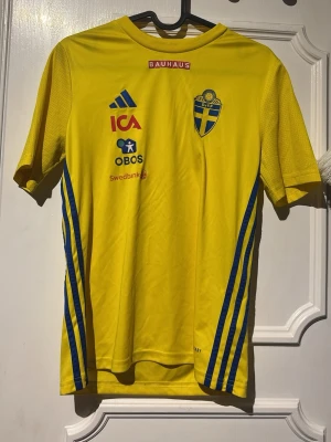 Svensk landslagströja Adidas gul - Gul kortärmad landslagströja från Adidas med blå detaljer och svenska landslagets emblem på bröstet. Tröjan har tryckta sponsorer som ICA, OBOS och Swedbank samt blå ränder längs sidorna. Perfekt för fotbollsträning eller att heja på Sverige.