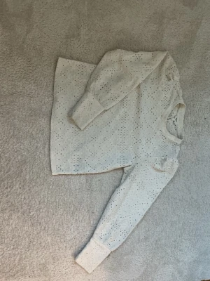 Vit broderad blus med lång ärm - Supersöt vit blus med broderade detaljer och hålmönster. Blusen har långa ärmar med breda muddar och rund halsringning. Perfekt för en fräsch och stilren look. Materialet känns lätt och luftigt.