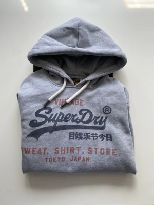 Grå Superdry hoodie med tryck - Snygg grå hoodie från Superdry med stor logga och text i svart och rött framtill. Klassisk huva med vita dragsnören och mjukt material som känns skönt mot huden. Perfekt för en avslappnad och cool stil. Storlek M. Använd ca 5 ggr 