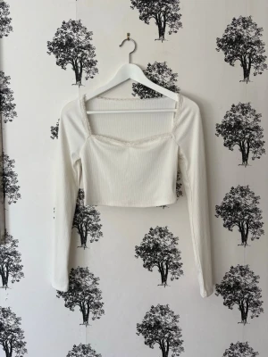 Vit croppad topp med spetsdetalj - Trendig vit croppad topp med långa ärmar och fyrkantig urringning. Toppen har fina spetsdetaljer längs halsringningen och är tillverkad i ett ribbat material som ger en snygg struktur. Perfekt att styla med höga jeans eller kjol.