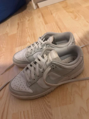 Vita Nike Dunk Low sneakers - Säljer ett par klassiska Nike Dunk Low sneakers i vitt med ljusgrå detaljer. Skorna har perforerad tå, snörning och den ikoniska swoosh-loggan på sidan. Perfekta för dig som gillar stilrena och tidlösa sneakers.
