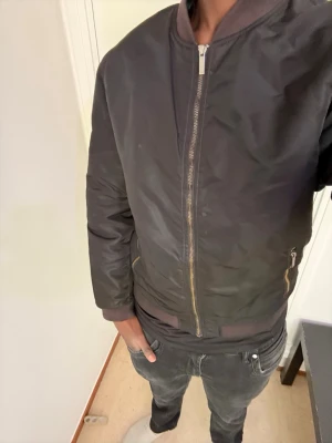 Svart bomberjacka från Esprit - Snygg svart bomberjacka från Esprit med klassisk ribbad krage, muddar och dragkedja framtill. Jackan har två fickor med dragkedja och är tillverkad i ett glansigt material som ger en modern look. Perfekt för dig som vill ha en stilren och enkel jacka.