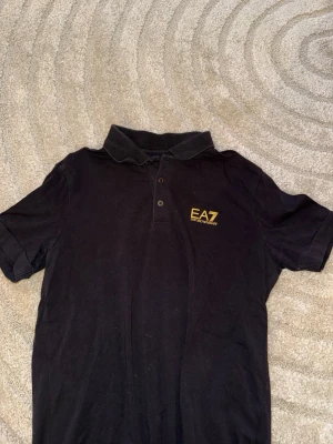 Svart pikétröja från EA7 Emporio Armani - Svart pikétröja från EA7 Emporio Armani med klassisk krage och knäppning framtill. Tröjan har korta ärmar och en gul EA7-logga på bröstet. Perfekt för en stilren och sportig look. Använd fåtal gånger. Super skön tröja. 
