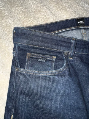 Mörkblå jeans från BOSS - Snygga mörkblå jeans från BOSS med klassisk femficksmodell och raka ben. Jeansen har diskreta kontrastsömmar och en liten BOSS-logga på myntfickan. Tillverkade i slitstark denim som ger en tidlös look. Super bra skick på dem och bra passform, är 180 cm. 