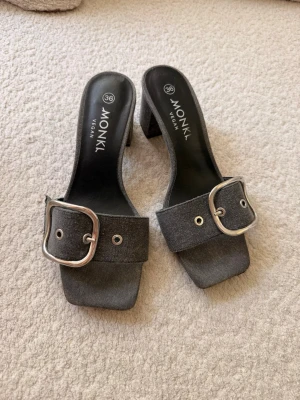 Gråa sandaler med klack från Monki - Snygga jeans gråa sandaler från Monki, bred rem och stor silverfärgad spänndetalj. Modellen har klack och är tillverkad i veganskt material. 