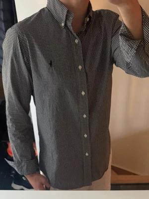Rutig skjorta från Ralph Lauren - Klassisk svartvit rutig skjorta från Ralph Lauren med button-down krage och broderad logga på bröstet. Skjortan har lång ärm, vita knappar och är tillverkad i mjuk bomull. Perfekt för en stilren och avslappnad look.