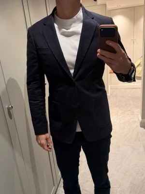 Marinblå kavaj från Tiger of Sweden  - Säljer en stilren marinblå kavaj med slimmad passform och snygga detaljer som knappar på ärmsluten och två framfickor. Kavajen har en bröstficka och är tillverkad i ett slätt material som ger ett elegant intryck. Perfekt för dig som vill ha en tidlös look. 
