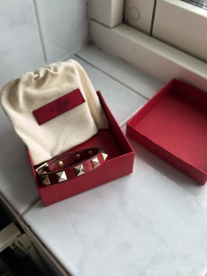 Valentino Armband röd. - Säljer ett snyggt rött armband från Valentino i äkta läder med ikoniska guldfärgade pyramidnitar. Armbandet har justerbart spänne och kommer i originalförpackning med tygpåse samt 2 extra nitar. Perfekt accessoar för att lyfta din outfit.
