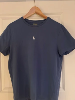 Polo Ralph Lauren t-shirt – blå – fint skick - Säljer en stilren blå t-shirt från Polo Ralph Lauren i custom slim fit. Klassisk modell med broderad logga – passar till allt. Säljer då den tyvärr är för liten för mig. Köpte för 600, säljer för 179kr Storlek: M                                                                           Skick: Mycket bra skick, inga synliga defekter