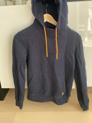 Mörkblå hoodie med orange snören från Pier One - Snygg mörkblå hoodie med huva och orangea dragsnören från Pier One. Hoodien har en stor magficka och ribbade muddar vid ärmslut och nederkant. Perfekt för en avslappnad stil. Materialet känns mjukt och bekvämt, troligen bomullsblandning.