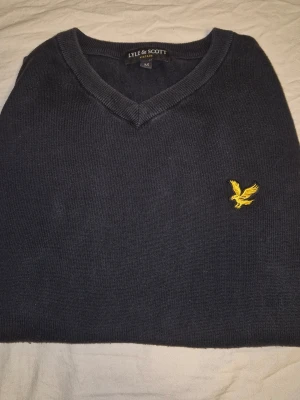 Svart stickad tröja Lyle & Scott - Produktdetaljer: Märke: Lyle & Scott Modell: Långärmad tröja med V-ringning Färg: Marinblå Passform: Regular fit Detalj: Broderad logotyp (gyllene örn) på bröstet Halsringning: V-ringad Ärmar: Långärmad Storlek: M (enligt etikett på bilden)