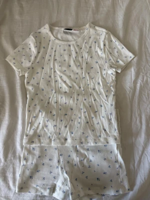 Blommigt ribbat set från Gina Tricot - Sött tvådelat set från Gina Tricot i ribbat material. Setet är vitt med små blå blommor och består av en kortärmad topp och shorts. Perfekt för en mysig dag hemma ellercatt sova i. Materialet är mjukt och stretchigt för extra komfort.