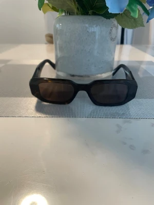 Bruna solglasögon Prada - Snygga svarta solglasögon från Prada med rektangulära bågar och diskret logga på sidan. Glasögonen har en stilren och modern look, perfekta för att lyfta din outfit. Bågarna är i plast och har en mörk ton på glasen.
