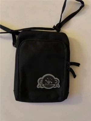 Svart axelväska med patch - Snygg svart axelväska i kompakt modell med justerbar axelrem och dragkedja. Väskan har en patch med sofialäktar motiv framtill och flera fack för småprylar. Perfekt för att bära det viktigaste när du är på språng.