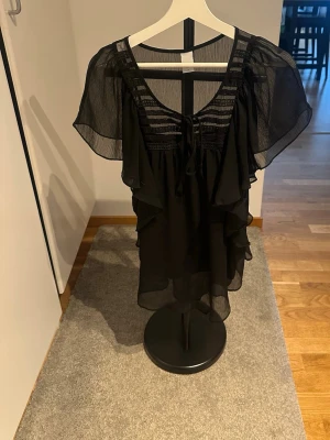 Svart transparent volangblus - Snygg svart blus i transparent material med volangdetaljer och korta, vida ärmar. Blusen har rund halsringning med knytband framtill och dekorativa ränder upptill. Perfekt för en trendig och luftig look.
