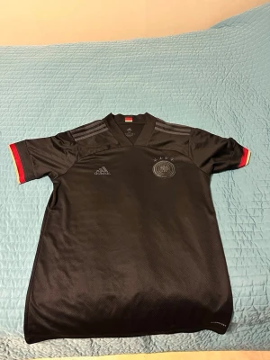 Svart Adidas fotbollströja Tyskland - Snygg svart fotbollströja från Adidas med Tysklands landslagslogga på bröstet och fyra stjärnor ovanför. Tröjan har röda och gula detaljer vid ärmsluten och klassiska Adidas-ränder på axlarna. Perfekt för dig som älskar fotboll och vill visa ditt stöd för Tyskland.