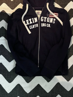 Marinblå Lexington zip-hoodie - Snygg marinblå hoodie från Lexington med dragkedja, vit textprint på bröstet och amerikansk flaggpatch. Utrustad med huva och vita dragsnören samt två fickor framtill. Perfekt för en avslappnad stil. Kostar 1500 ny och i väldigt fint skick. Funderingar? Tveka inte på att höra av dig mvh Strwwt wst☺️