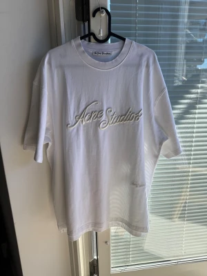 Vit Acne Studios t-shirt med broderi - Vit t-shirt från Acne Studios med broderad logga framtill i vitt. Klassisk passform med rund halsringning och korta ärmar. Tillverkad i mjuk bomull, perfekt för en clean och stilren look.