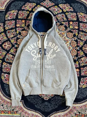Grå zip-hoodie från Tokyo Laundry - Snygg grå hoodie med dragkedja från Tokyo Laundry. Stor vit print framtill med text och årtal, samt kängurufickor och justerbar huva med vita snören. Mörkblått innerfoder i huvan ger en schysst kontrast. Perfekt för en avslappnad streetstil.