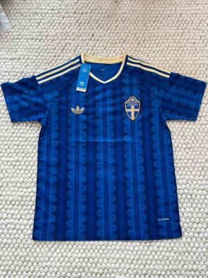 Blå Sverige fotbollströja Adidas - Snygg blå fotbollströja från Adidas med Sveriges landslagsemblem på bröstet och klassiska gula detaljer. Tröjan har ett mönster i olika blå nyanser, tre gula ränder på axlarna och gul krage. Tillverkad i lätt och funktionellt material.