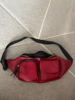 Röd magväska med svarta detaljer - Cool röd magväska i slätt material med två framfickor, en med dragkedja och en med lock. Justerbart svart band och dragkedja upptill. Perfekt för att bära det viktigaste och ge outfiten en färgklick.