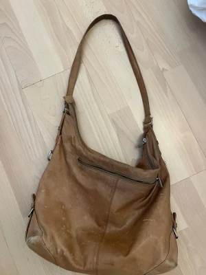 Brun axelväska i äkta läder - Snygg brun axelväska i äkta läder med flera fack, både utvändigt och invändigt. Väskan har en avslappnad, slouchy form och stängs med dragkedja. Perfekt för dig som vill ha plats för allt du behöver under dagen.