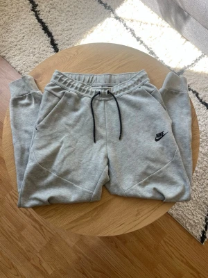 Gråa Nike Tech mjukis - Hej👋 Säljer nu dessa snygga gråa mjukisbyxor från Nike med svart dragsko i midjan och klassisk Nike-logga på benet🤩 Byxorna har två sidofickor och är tillverkade i mjukt och bekvämt material, perfekt för chill dagar eller träning👌 Vid frågor meddela gärna📩