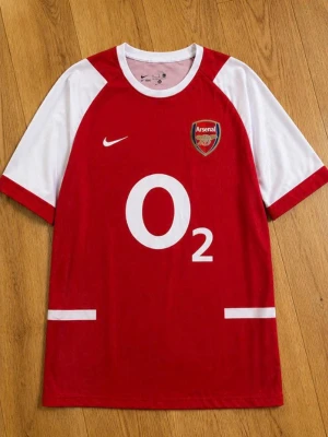 Arsenal Bergkamp 10 matchtröja Nike - Klassisk röd och vit Arsenal matchtröja från Nike med O2 sponsortryck, klubbmärke på bröstet och Bergkamp 10 på ryggen. Tröjan har korta ärmar och är tillverkad i lätt och ventilerande material, perfekt för fotboll eller samling.