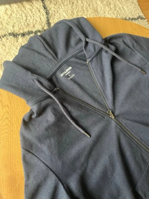 Mörkblå zip-hoodie från Jack & Jones - Tjena👋 Säljer en mörkblå hoodie från Jack & Jones med dragkedja framtill och justerbar huva med snören🤩 Hoodien har långa ärmar och ribbade muddar vid ärmslut och nederkant👍 Perfekt för en avslappnad och stilren look👌 meddela gärna vid frågor📩