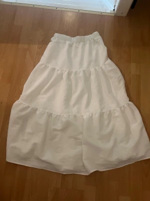 Vit volangkjol med resårmidja - Säljer en lång, vit kjol med flera volanger och resår i midjan. Kjolen har en luftig och flowy känsla, perfekt för varmare dagar. Materialet känns mjukt och lätt, vilket gör den superbekväm att bära.