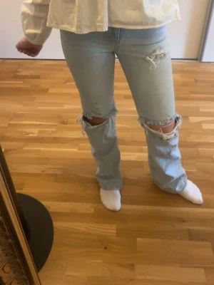 Ljusblå ripped jeans med raka ben - Säljer ett par ljusblå jeans med raka ben och stora slitningar vid knäna. Jeansen har en avslappnad passform och klassisk femficksdesign. Perfekta för en trendig och avslappnad stil. Materialet är denim i bomull.