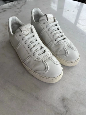 Vita sneakers från Valentino - Snygga vita sneakers från Valentino i läder med klassisk låg profil och diskreta detaljer. Skorna har vita snören, mönstrad sula och logotyp på plösen. Perfekta för en clean och stilren look.