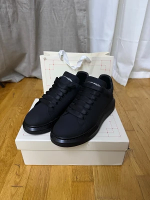  Alexander McQueen  - Stilrena svarta sneakers från Alexander McQueen med chunky sula och breda svarta skosnören. Skorna har en låg siluett och är tillverkade i slätt läder med logga på plösen. Perfekta för dig som gillar minimalistisk och modern design.