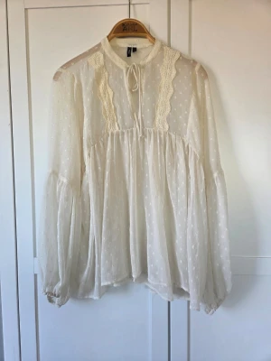 Vit transparent blus med spetsdetaljer - Romantisk vit blus från Vero Moda i transparent, prickigt tyg med spetsdetaljer längs bröstet och knytband vid halsen. Blusen har långa, puffiga ärmar med resår vid ärmslut och en lös, flowy passform. Perfekt för en bohemisk look.