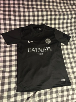 Svart PSG x Balmain t-shirt Nike - Svart t-shirt med Paris Saint-Germain och Balmain Paris-logga på bröstet. Klassisk rund hals och korta ärmar. Tillverkad i ett lätt och sportigt material, perfekt för träning eller streetwear. Snygg vit Nike swoosh på framsidan.