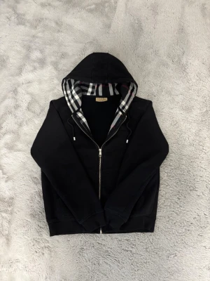 Svart hoodie med dragkedja Burberry - Svart hoodie från Burberry med dragkedja och klassiskt rutigt foder i huvan och insidan. Tröjan har justerbar huva med snören och ribbade muddar. Perfekt för dig som gillar stilrena och exklusiva plagg med en twist.