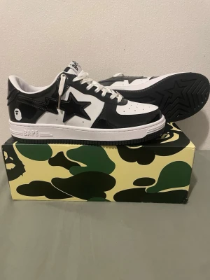 Svarta och vita BAPE STA sneakers - Snygga BAPE STA sneakers i svart och vitt läder med ikonisk stjärnlogga på sidan och BAPE-detaljer på hälen. Klassisk låg modell med perforerad tå och vita skosnören. Kommer i originalkartong med camomönster.