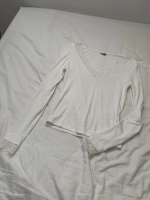 Vit långärmad topp med spetsdetaljer - Supersöt vit långärmad topp med v-ringning och spetsdetaljer vid halsringning och ärmslut. Perfekt att styla med jeans eller kjol för en fräsch look. Toppens material känns mjukt och lätt mot huden.