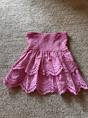Rosa bandeau topp med volang - Supersöt rosa bandeau topp med broderade volanger och hålmönster. Toppens överdel är smockad för en bekväm passform och den är axelbandslös. Perfekt för varma dagar och ger en gullig och trendig look.
