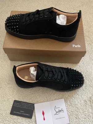 Christian Louboutin svarta sneakers - Svarta sneakers från Christian Louboutin i mocka med mattsvarta nitar på tån. Klassisk låg modell med svarta skosnören och diskret logga på insidan. Snygg och edgy design som sticker ut med sina detaljer. Levereras med originalkartong och tillbehör.