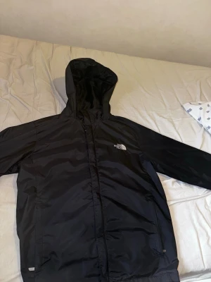 Svart vindjacka från The North Face - Snygg svart vindjacka från The North Face med huva och dragkedja framtill. Jackan har två sidofickor och logga broderad på bröstet. Tillverkad i lätt och tåligt material som skyddar mot vind och lätt regn. Perfekt för utomhusaktiviteter.
