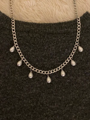 Silverfärgat halsband med pärldroppar - Stilrent halsband i silverfärgad rostfritt med sju hängande droppformade pärlor. Kedjan är grov och ger ett trendigt intryck, perfekt för att lyfta en enkel outfit. Pärlorna är vita och ger en elegant touch till halsbandet.