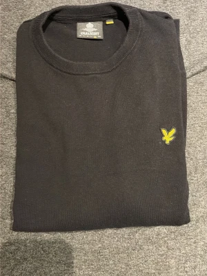 Svart stickad tröja från  Lyle & Scott - Svart rundhalsad tröja från Lyle & Scott med den klassiska gula örnloggan broderad på bröstet. Tröjan har ribbade muddar vid hals, ärmslut och nederkant. Perfekt för en stilren och enkel look.