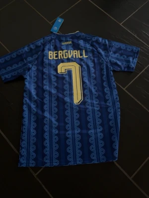 Blå Sverige fotbollströja Adidas - Säljer en blå Sverige fotbollströja från Adidas med gult tryck, nummer 7 och namnet Bergvall på ryggen. Tröjan har korta ärmar, gula ränder på axlarna och Sveriges landslagsemblem på bröstet. Tillverkad i lätt och ventilerande material.