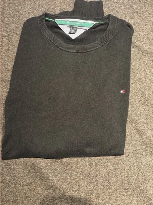 Svart stickad tröja Tommy Hilfiger - Klassisk svart stickad tröja från Tommy Hilfiger med rund halsringning och diskret logga på bröstet. Tröjan har ribbade muddar och är perfekt för en stilren look. Materialet är mjukt och bekvämt.
