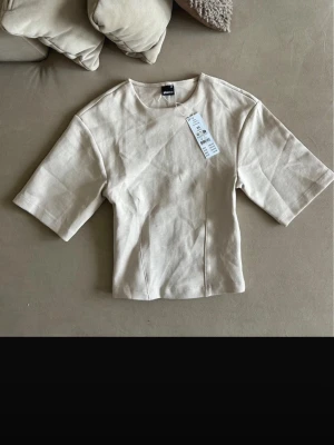 Beige topp från Gina Tricot - En stilren beige topp från Gina Tricot med rund halsringning och korta ärmar. Toppen har en rak passform och är tillverkad i mjuk bomull, perfekt för att skapa en clean och enkel look.