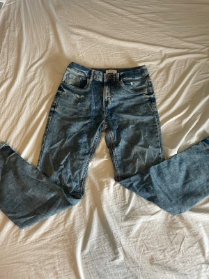 Blå jeans från Lindex med slitningar - Säljer ett par blå jeans från Lindex med coola slitningar och klassisk femficksdesign. Jeansen har en rak passform och är gjorda i denim med snygg tvättad look. Perfekta för dig som gillar en avslappnad och trendig stil.
